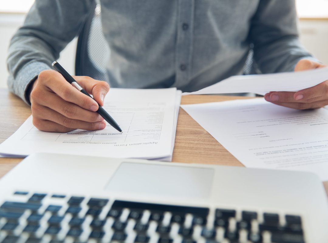 Unrecognizable businessman sitting at table holding ballpoint pen and papers and checking documents in office